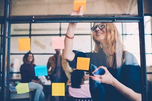 Smiling young female employee in stylish spectacles using stickers for creation strategy